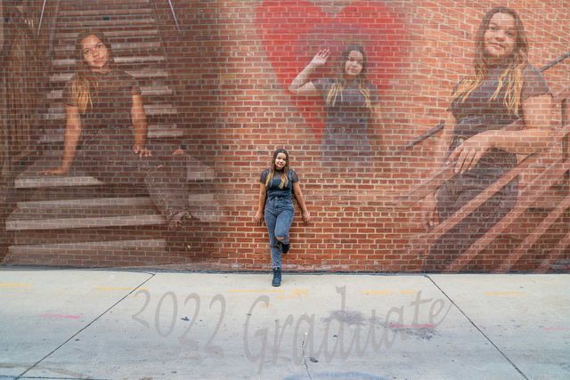 A woman is standing in front of a brick wall with a mural of three women.