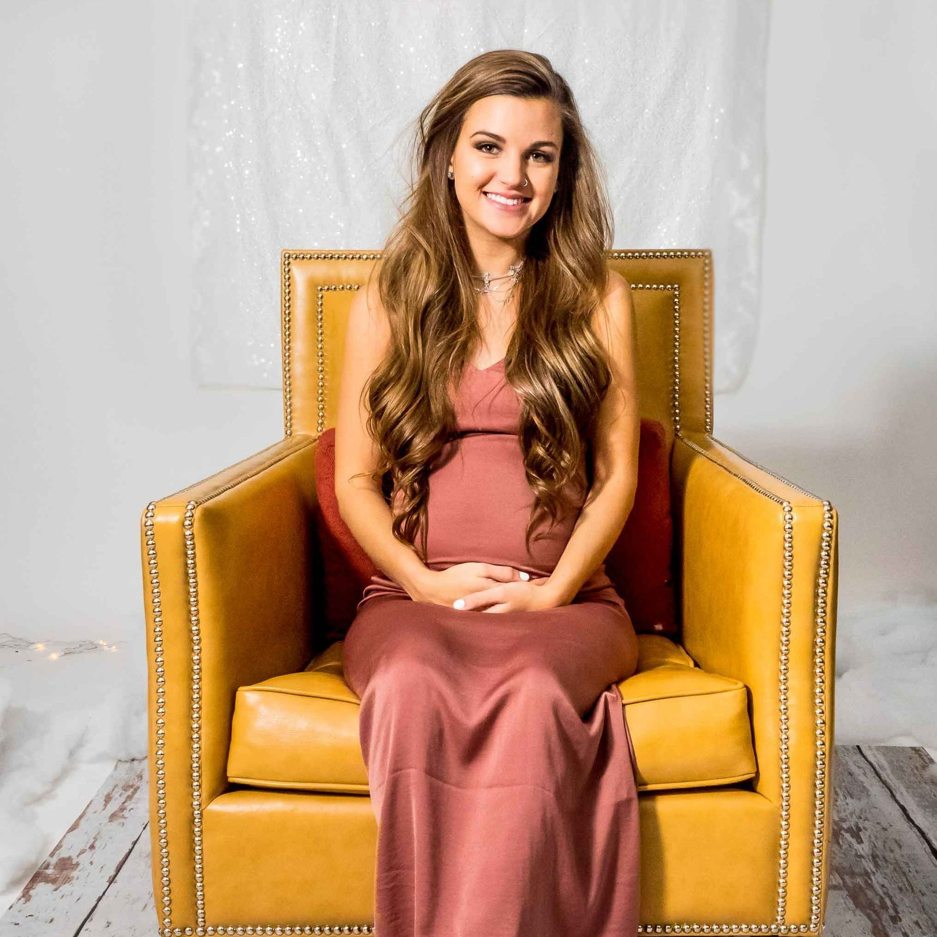 A pregnant woman in a pink dress is sitting in a yellow chair