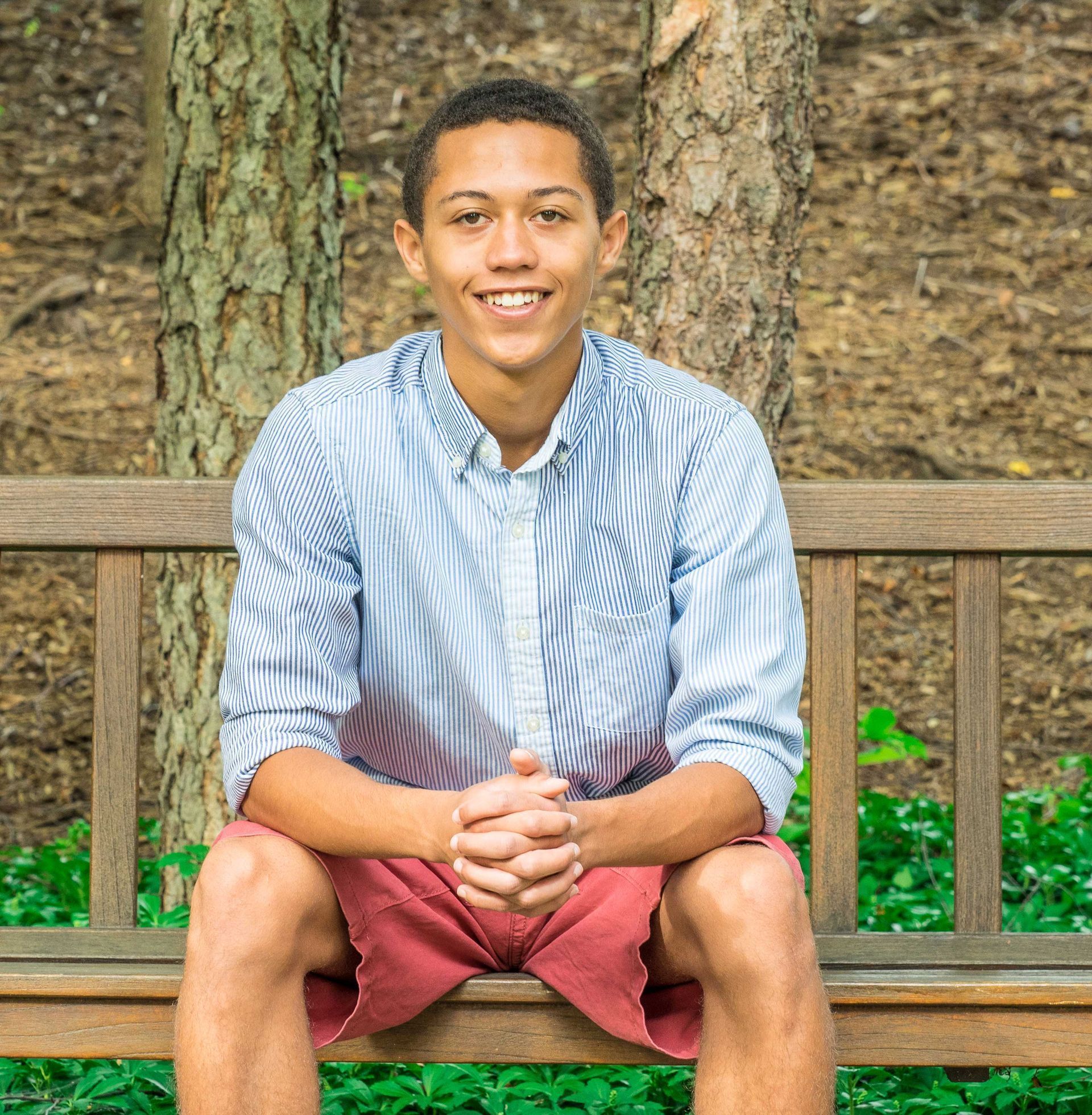 A young man is sitting on a wooden bench with his legs crossed and smiling.