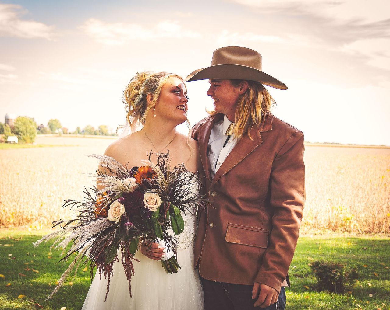 A bride and groom are posing for a picture in a field . the bride is wearing a cowboy hat.
