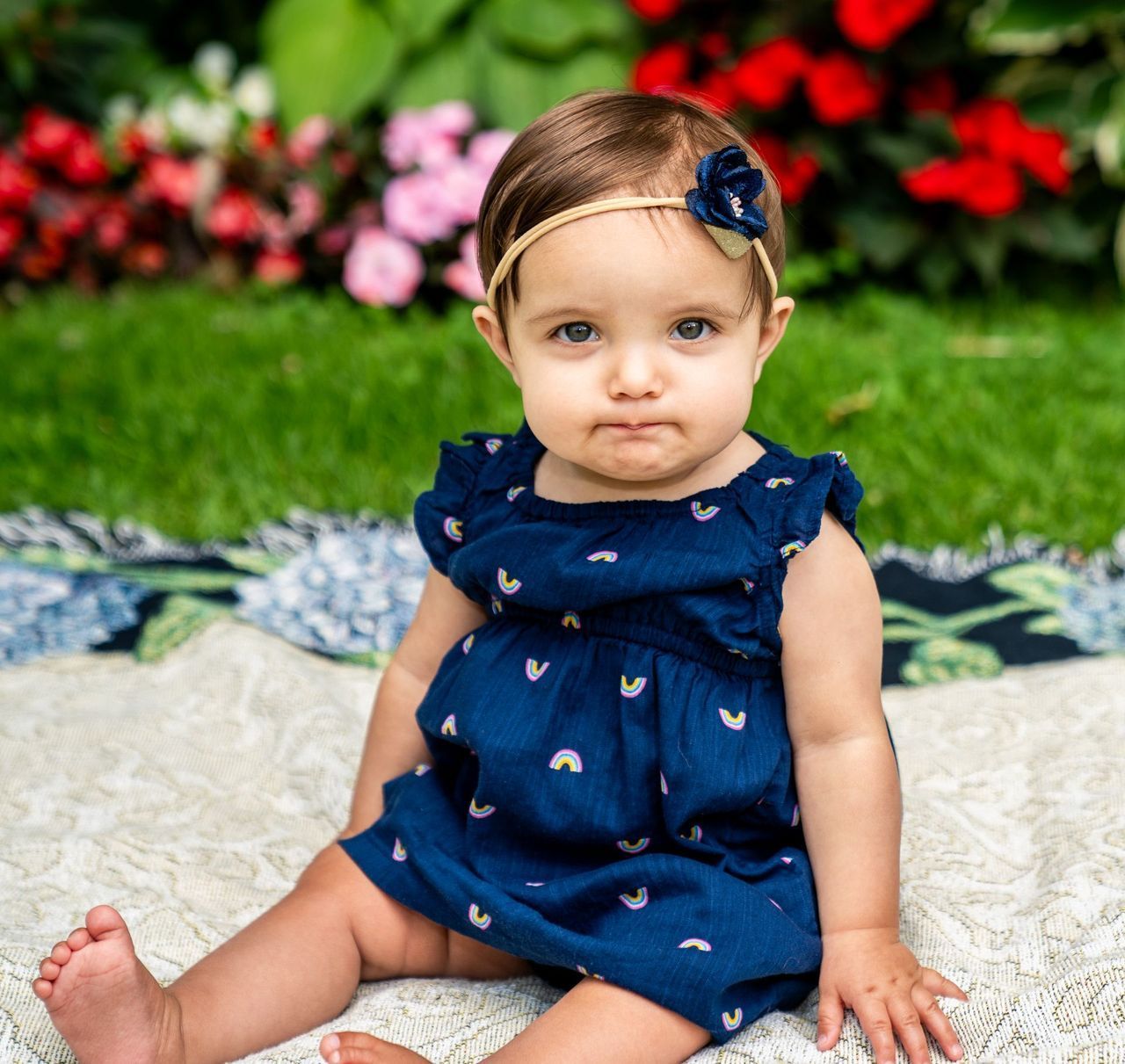 A baby girl in a blue dress is sitting on a blanket