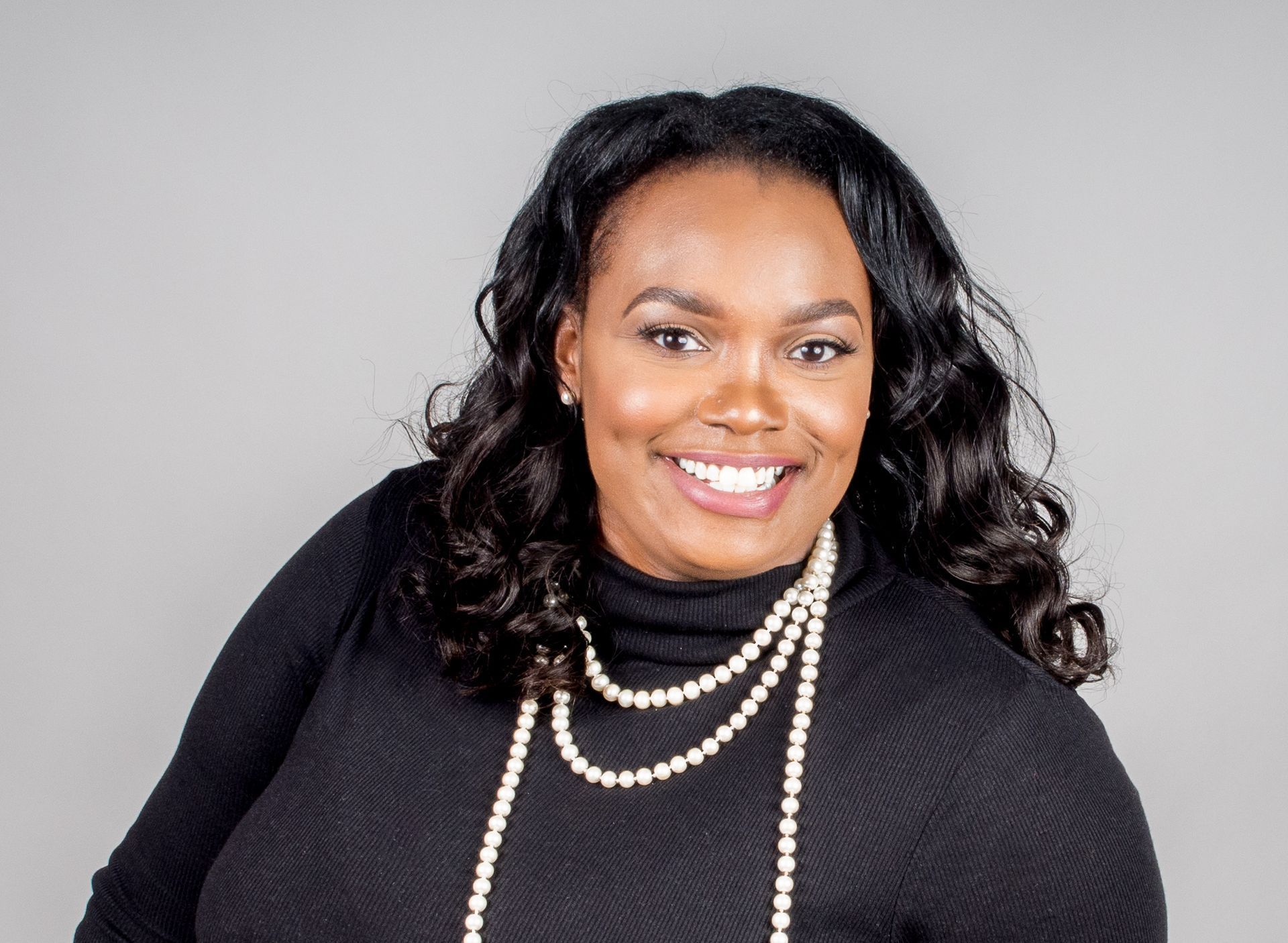 A woman wearing a black sweater and a pearl necklace is smiling.