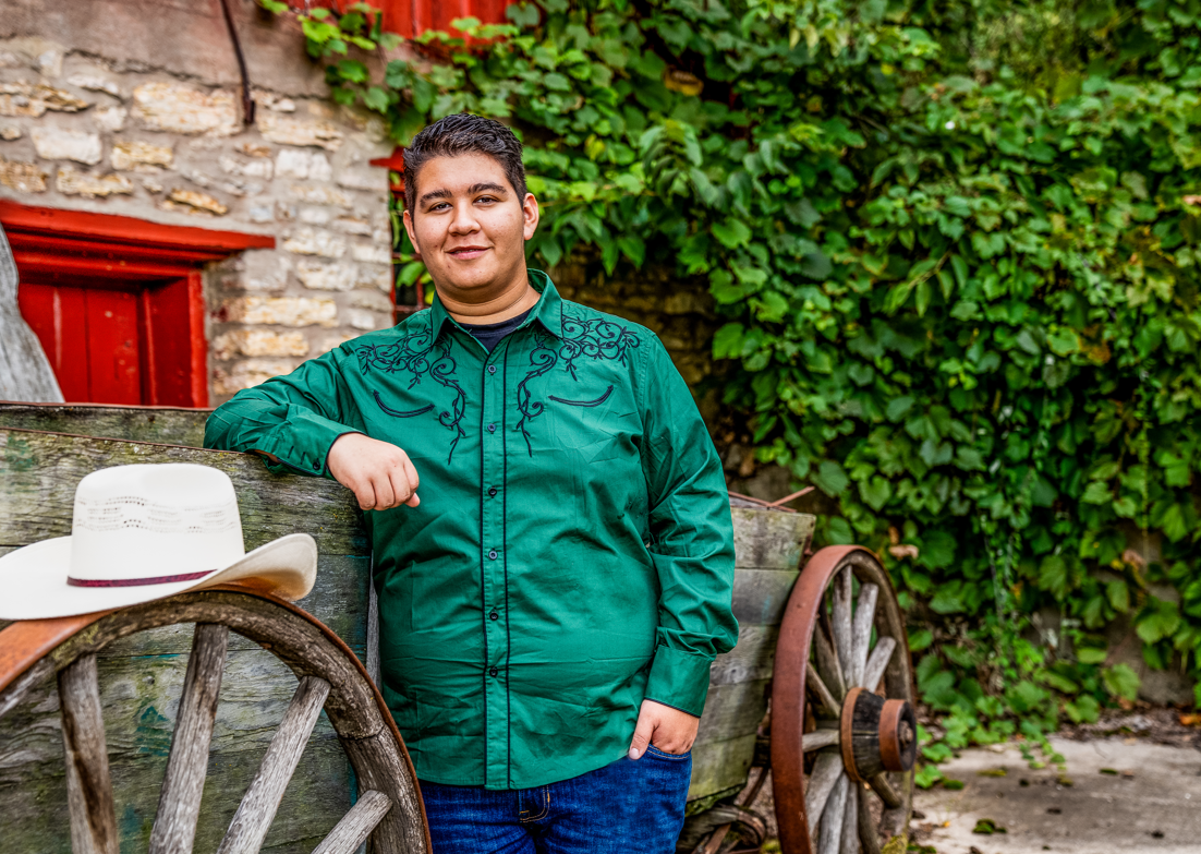 A man in a green shirt is leaning against a wall next to a wagon wheel and a cowboy hat.