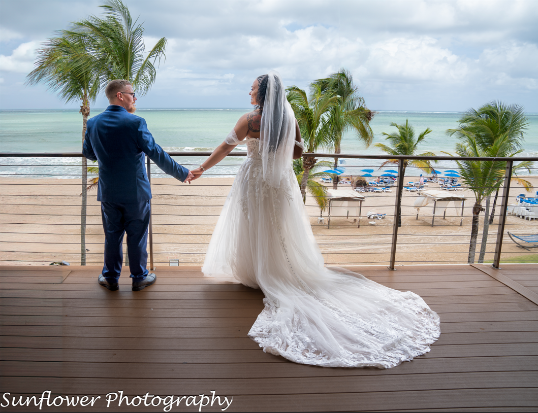 A bride and groom are holding hands on a balcony overlooking the ocean.