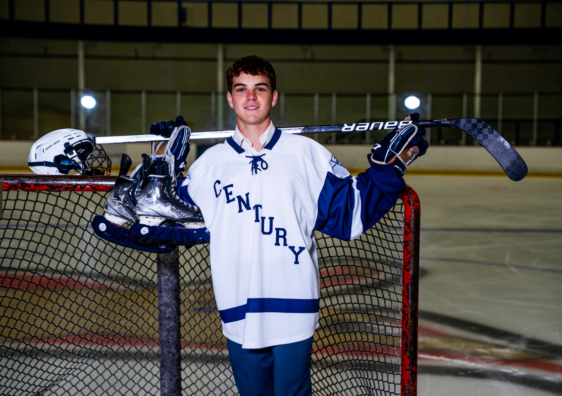 A young man in a century jersey is holding a hockey stick and ice skates.