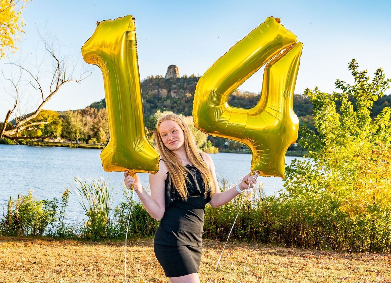 A woman in a black dress is holding a large gold balloon in the shape of the number 14.