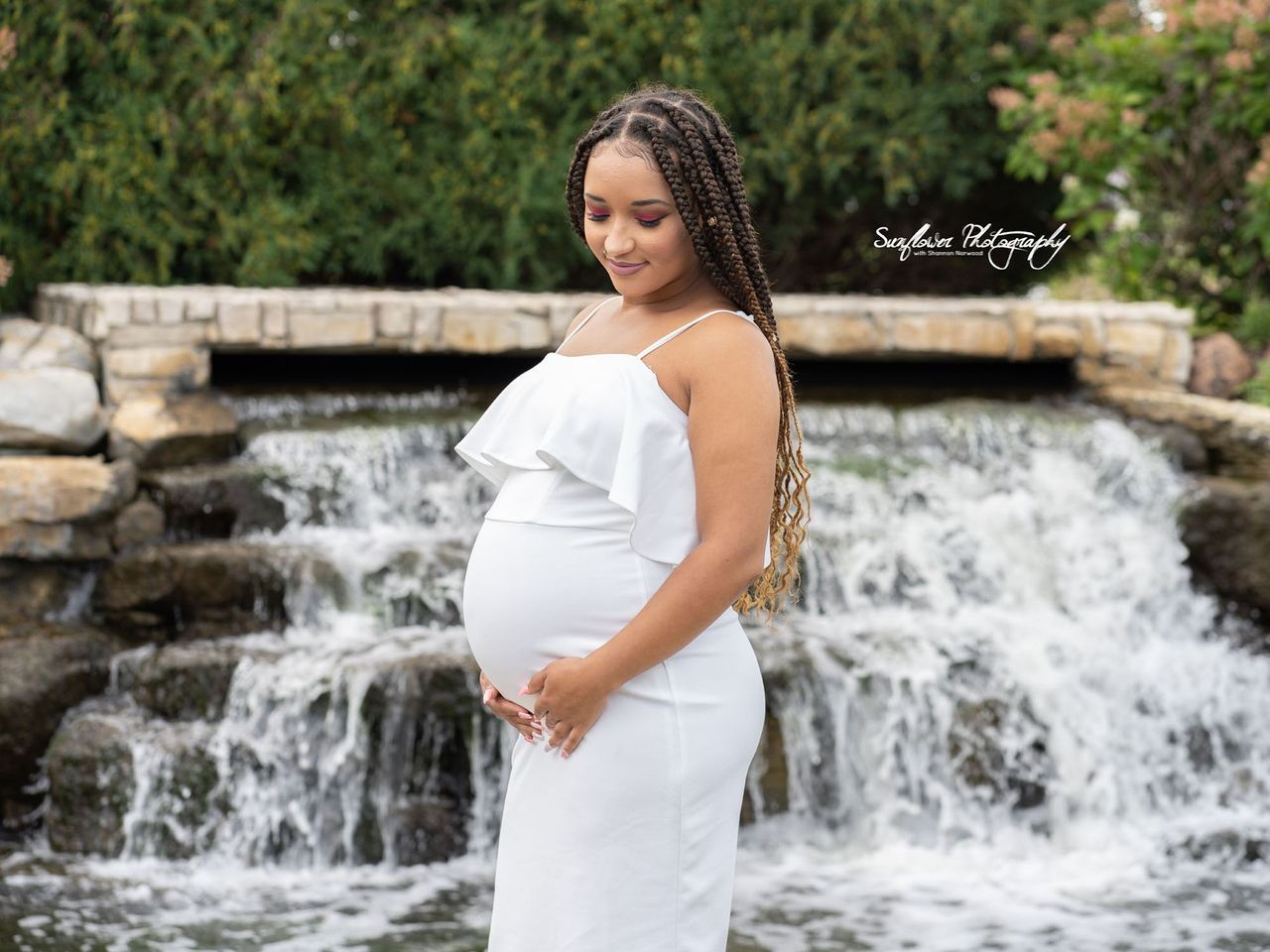 A pregnant woman in a white dress is standing in front of a waterfall.