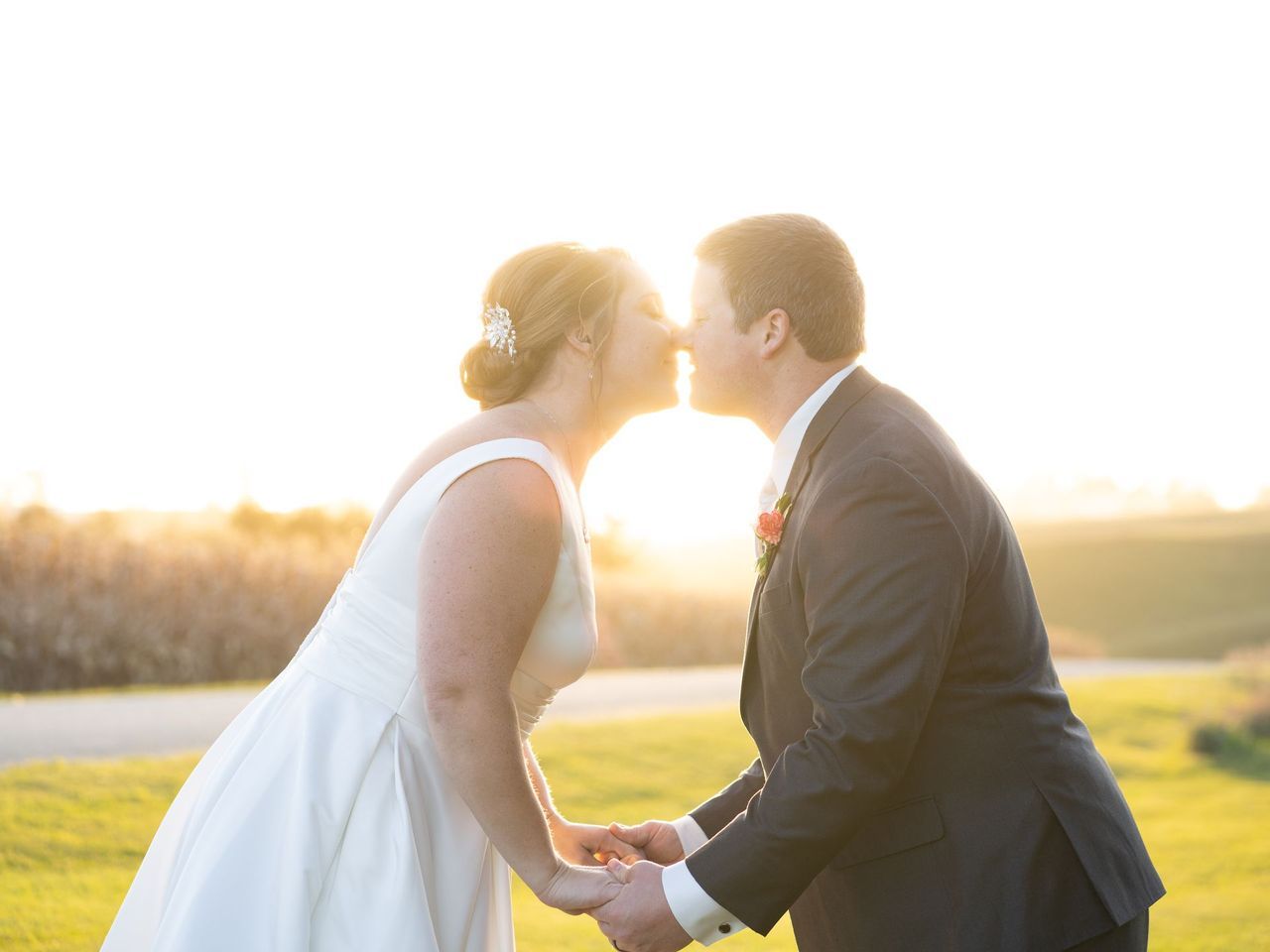A bride and groom are kissing in front of the sun while holding hands.