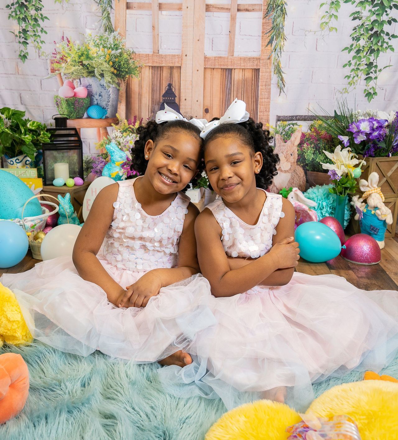 Two little girls are sitting next to each other on a rug surrounded by easter eggs.