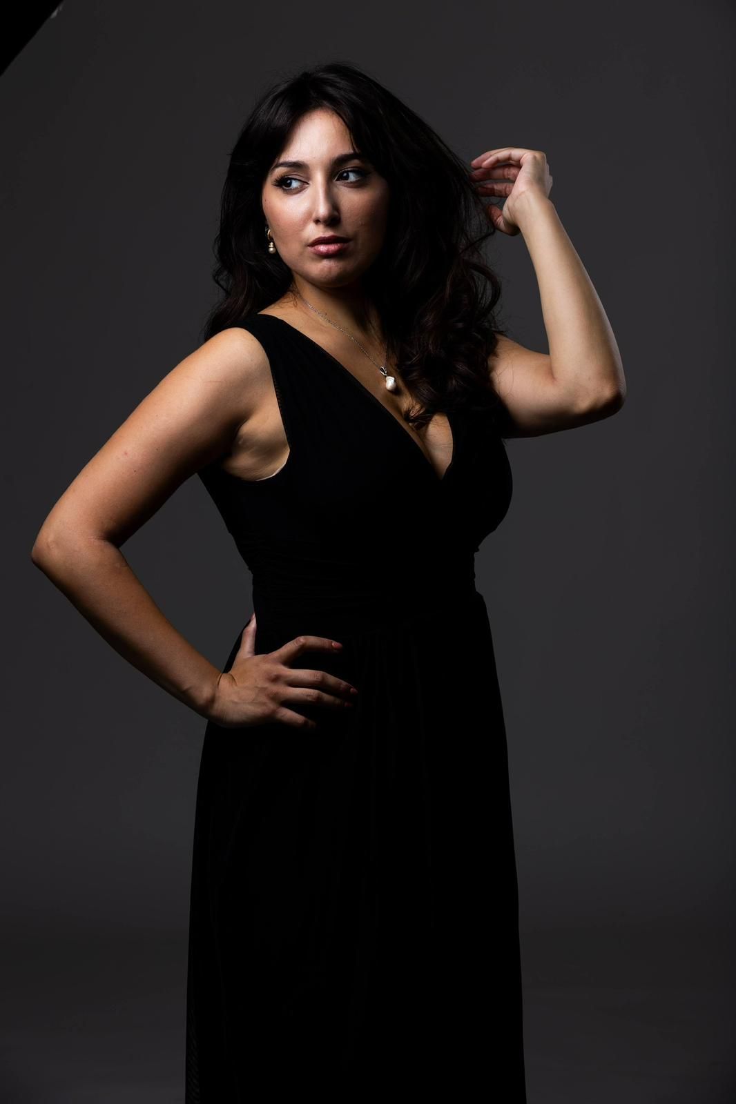 Donna in abito nero, mano sul fianco, tocca i capelli, sguardo rivolto a sinistra, sfondo grigio.