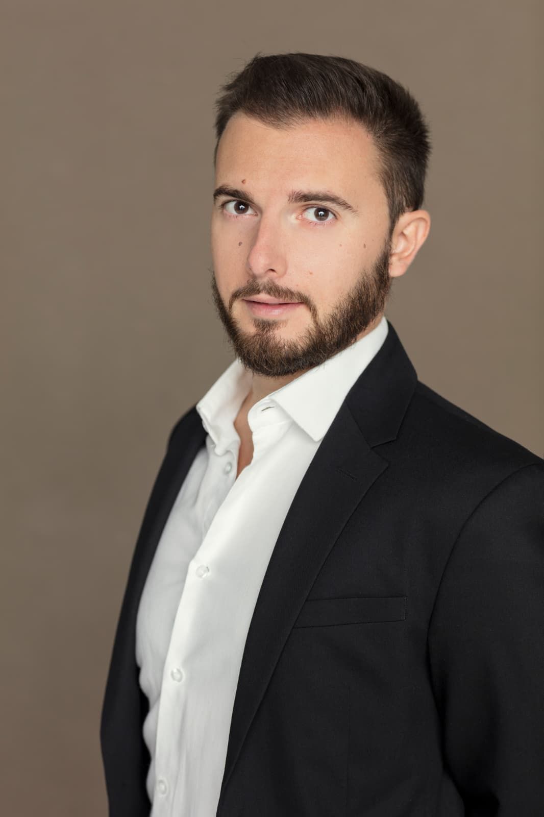 Uomo in abito nero e camicia bianca aperta, che guarda la telecamera. Sfondo neutro.