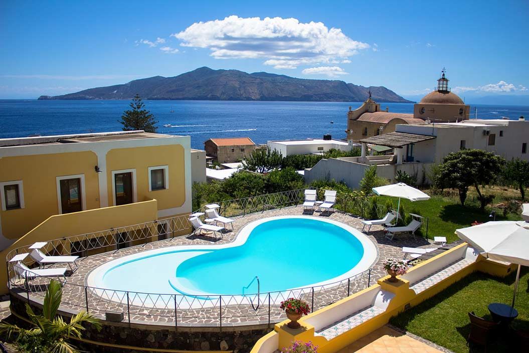 Piscina con vista sul mare, con una chiesa e un'isola sullo sfondo, sotto un cielo azzurro brillante.
