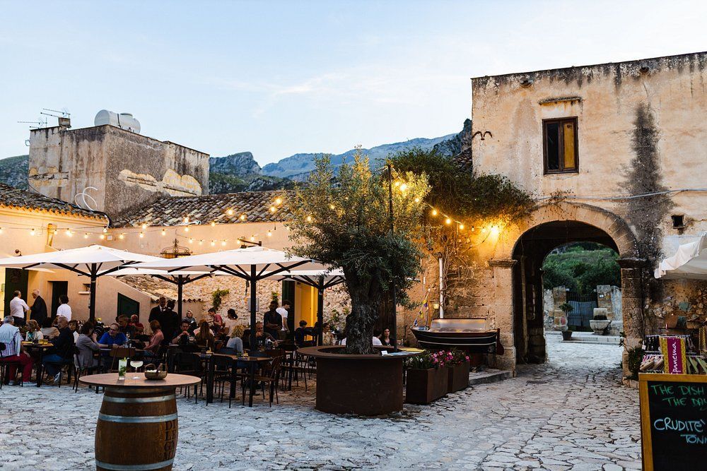Ristorante all'aperto al tramonto con persone, tavoli, ombrelloni e luci a ghirlanda in un cortile in pietra.