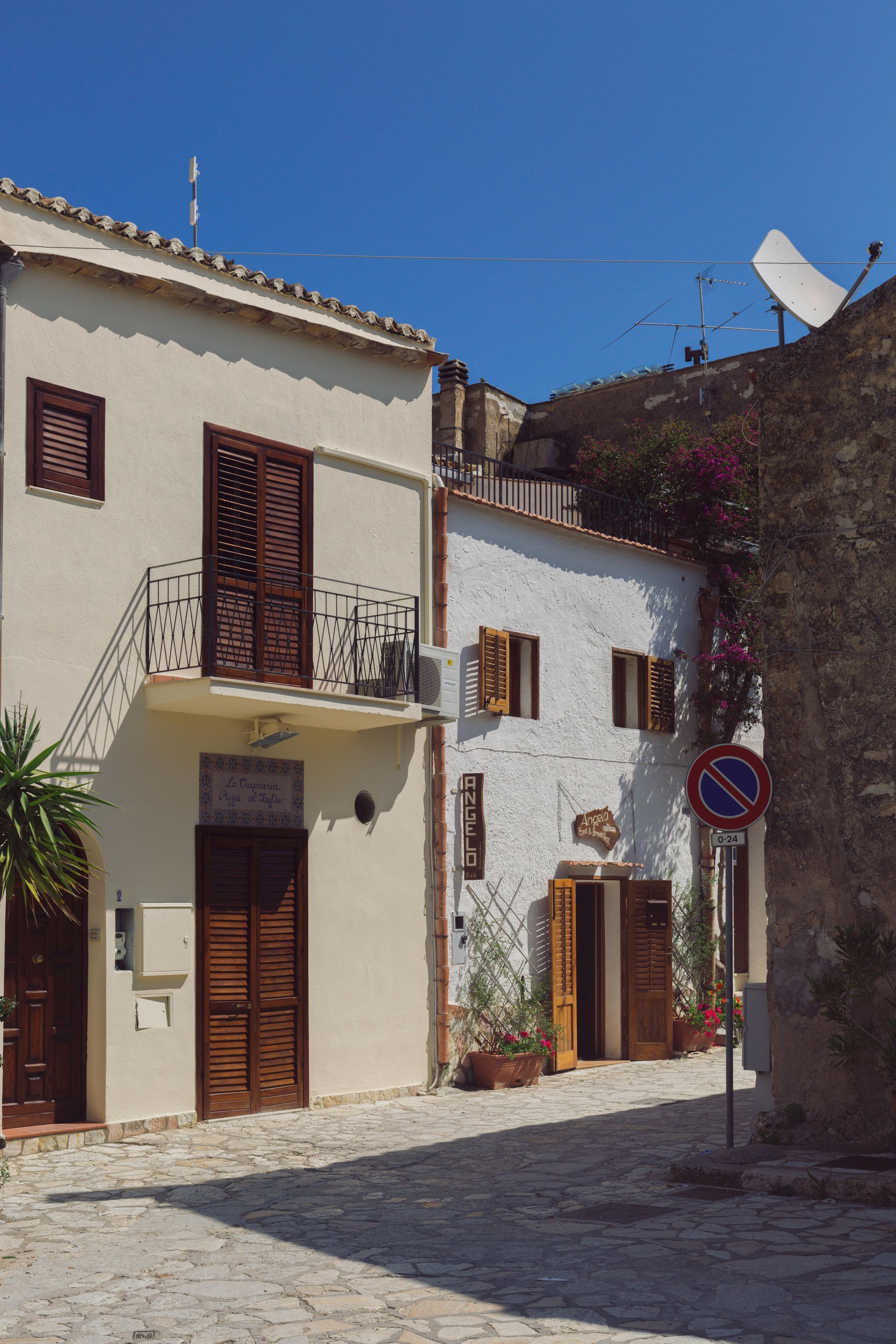 Strada lastricata in un villaggio italiano con edifici imbiancati, persiane marroni e cielo azzurro.