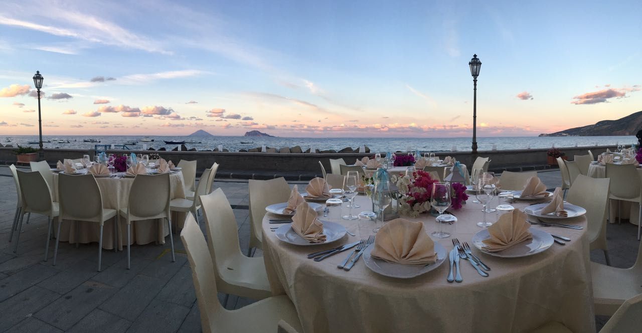 Tavoli all'aperto apparecchiati per una festa con vista sul mare al tramonto.
