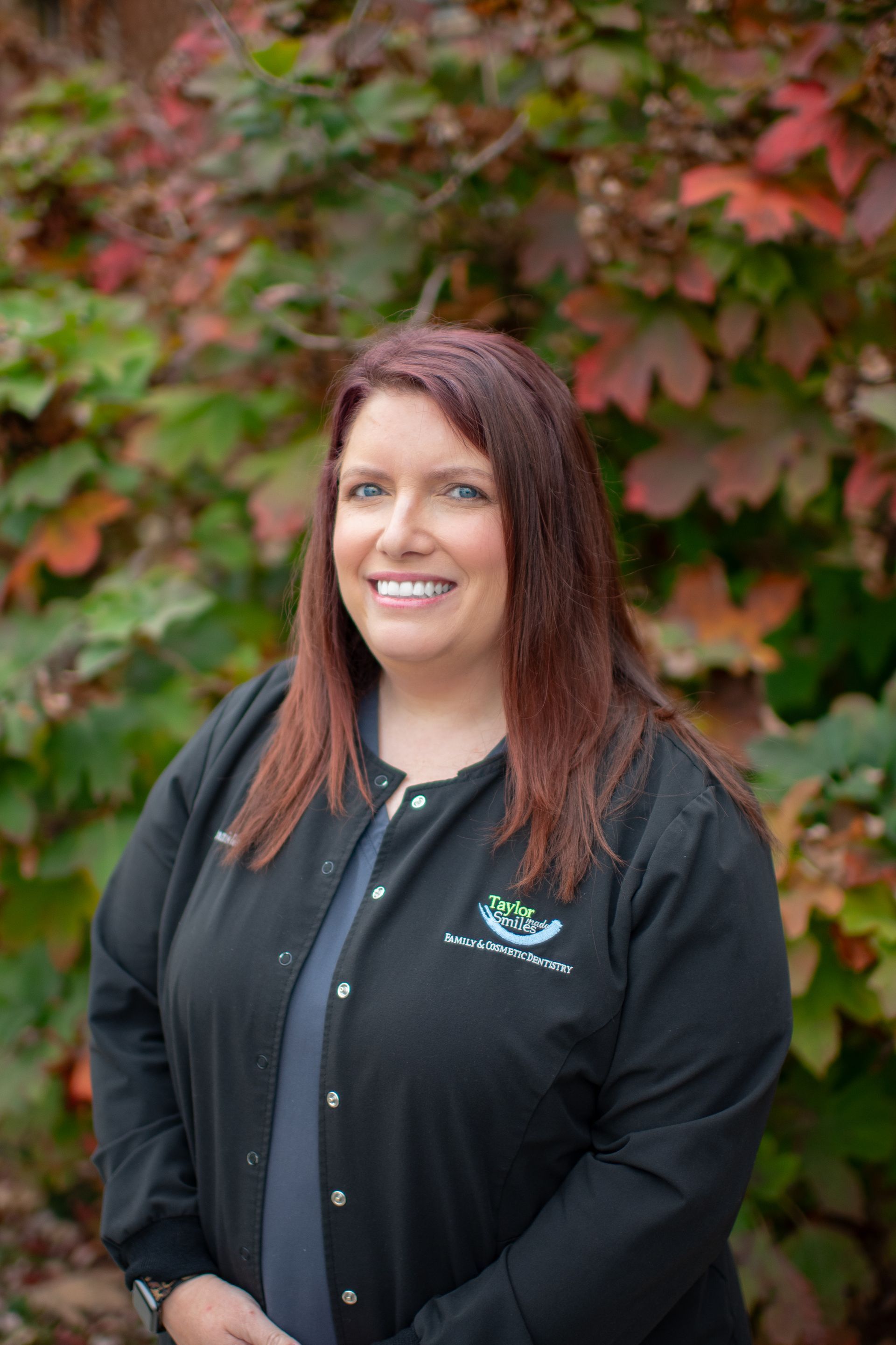 A woman in a black jacket is smiling in front of a bush.