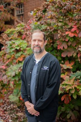 A man in a black jacket is standing in front of a bush.