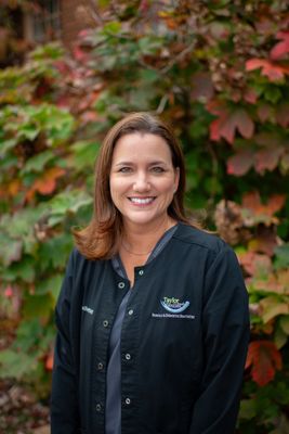 A woman in a black jacket is standing in front of a bush with red leaves.