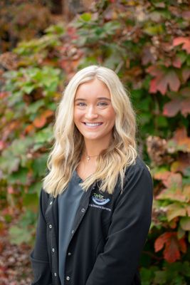 A woman in a black jacket is smiling in front of a bush.