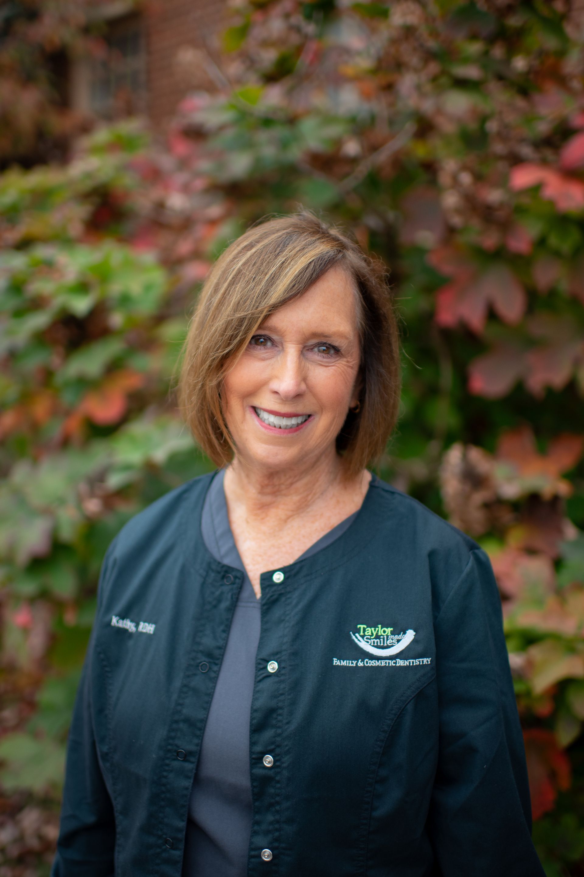A woman in a green jacket is smiling in front of a bush.