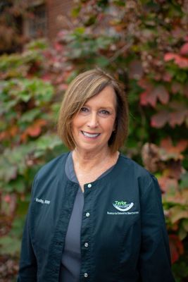 A woman in a green jacket is smiling in front of a bush.