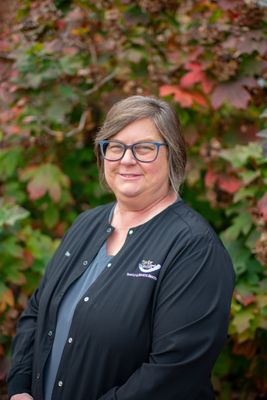 A woman wearing glasses and a black jacket is standing in front of a bush.