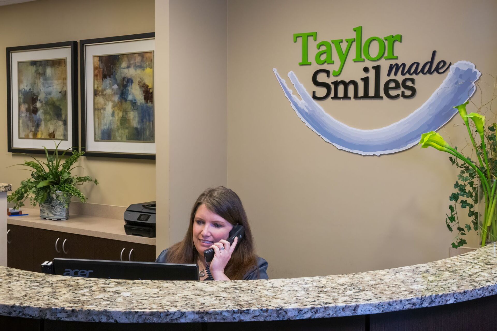 A woman is sitting at a desk in a dental office talking on a cell phone.
