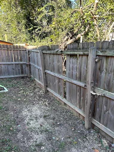 Damaged wooden fence in a backyard, missing pickets and boards, weathered and gray.