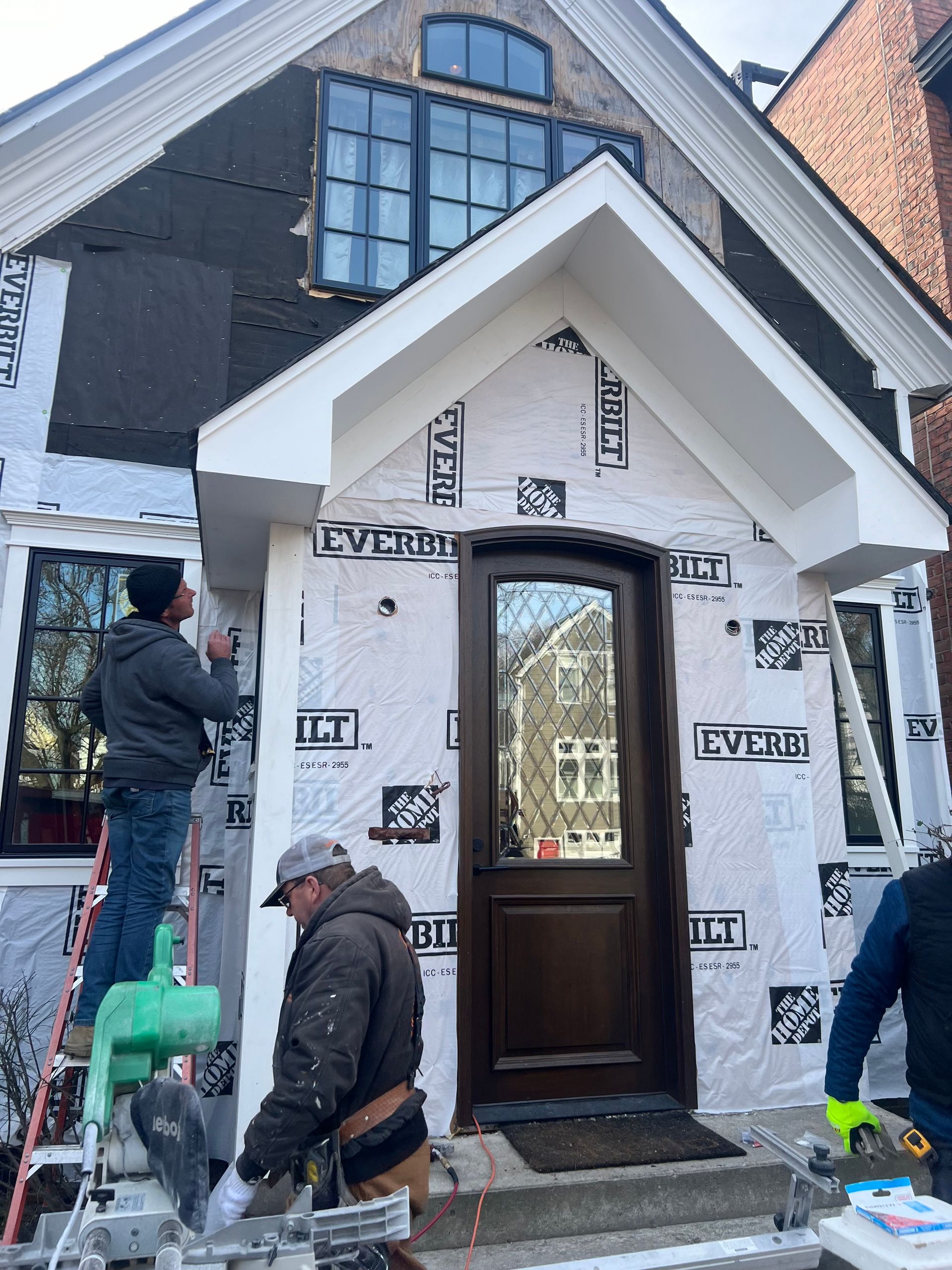 Construction workers installing trim on a house exterior. White siding, brown door, and black windows.
