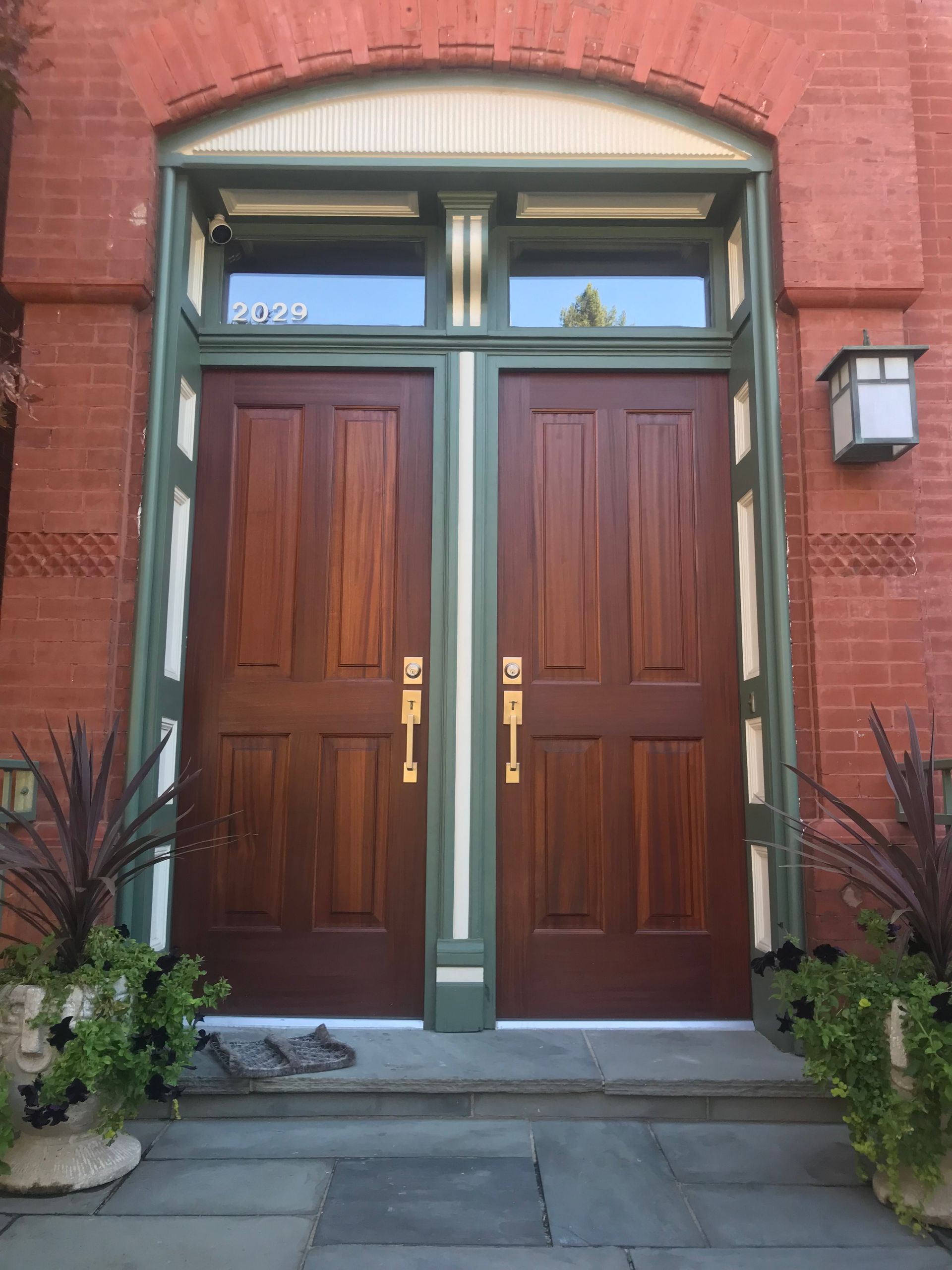 Double wooden doors with brass handles, set in a brick facade, green trim. Above, two small windows.