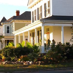 A white and pale green Victorian-style house with a long front porch, surrounded by lush landscaping and a stone border.