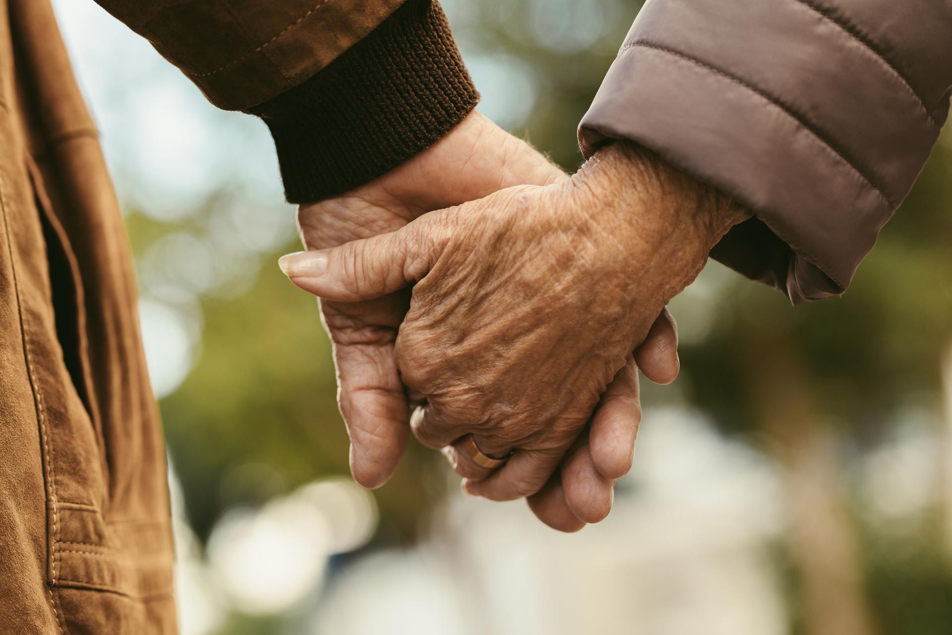 Two individuals hold hands, their fingers interlaced, set against a softly blurred natural outdoor background.