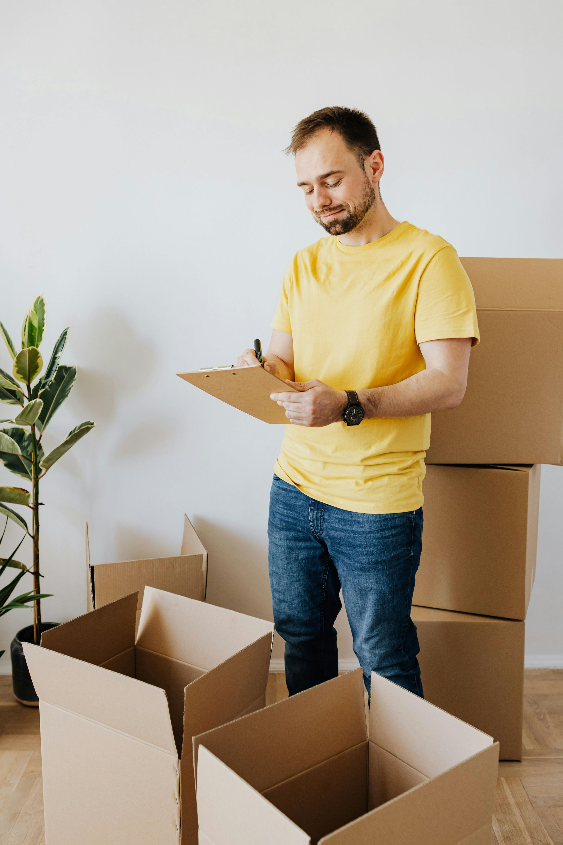 A person in a yellow shirt and jeans writes on a clipboard while standing among cardboard moving boxes in a room.