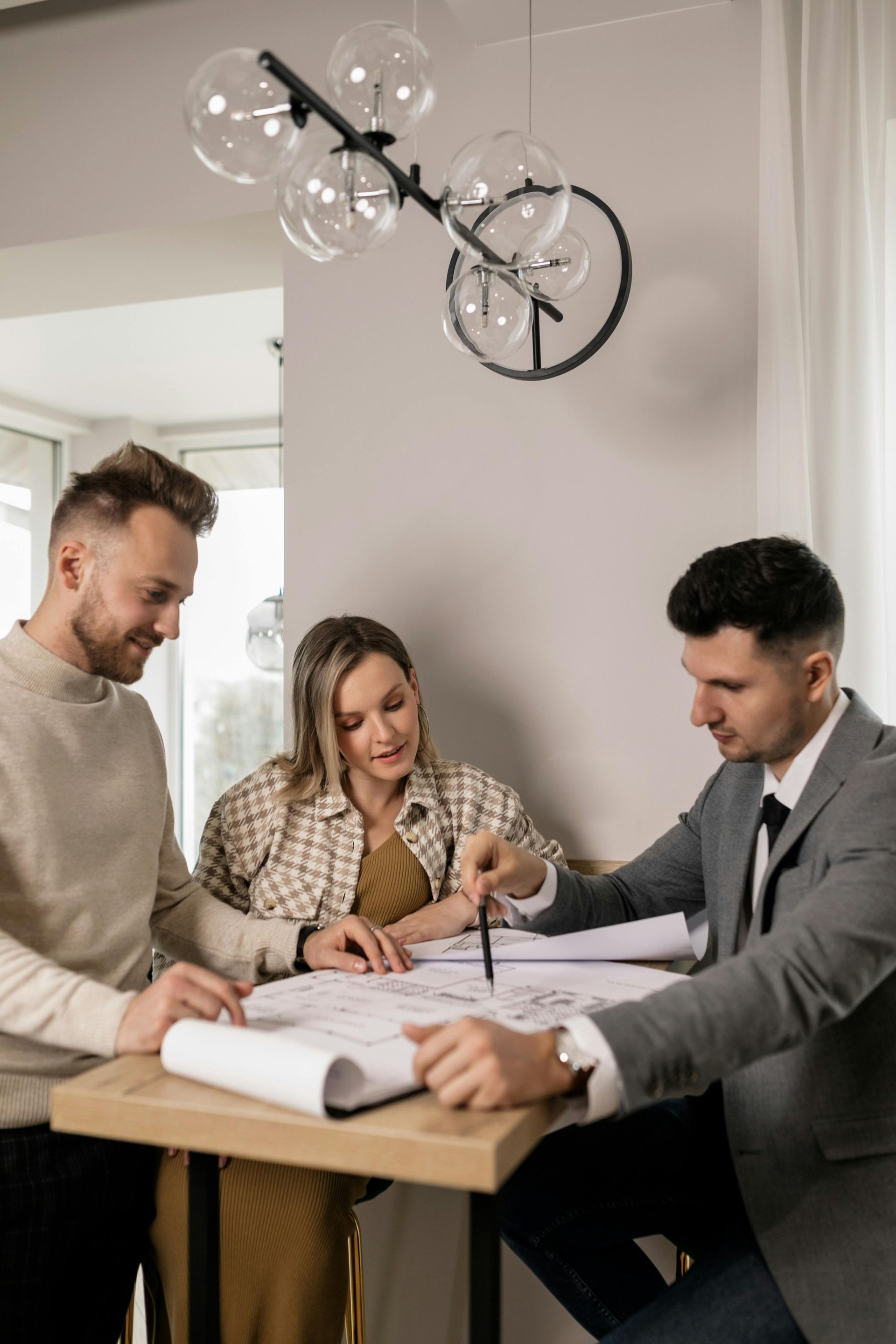 A professional in a suit reviews blueprints on a table with two individuals in a modern, well-lit interior.