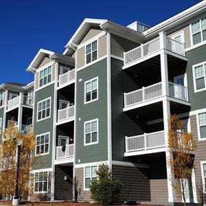 A multi-story apartment building with sage green siding, white balconies, and gabled roofs under a clear blue sky.