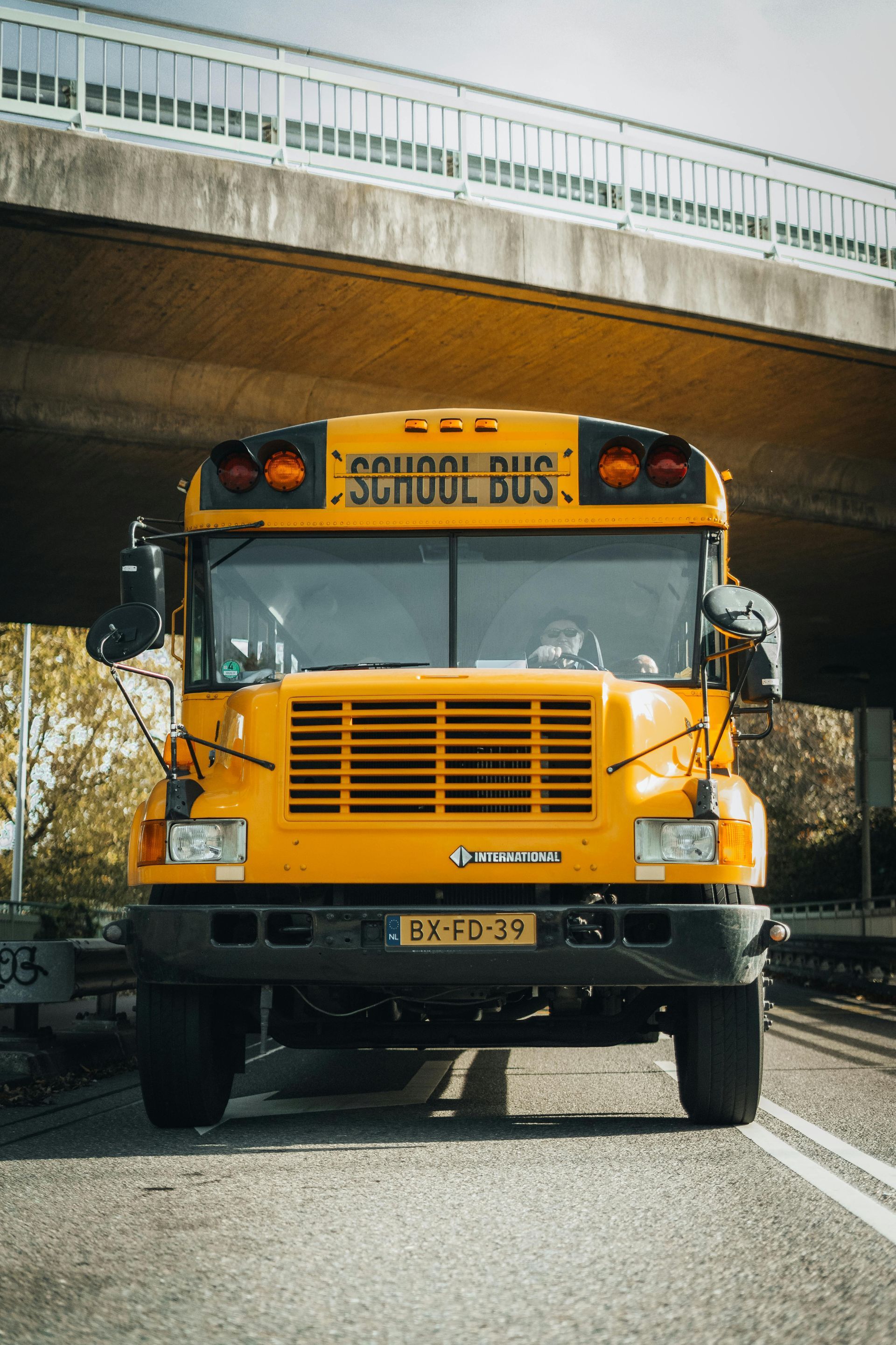 Yellow school bus driving on a road, under a concrete overpass.