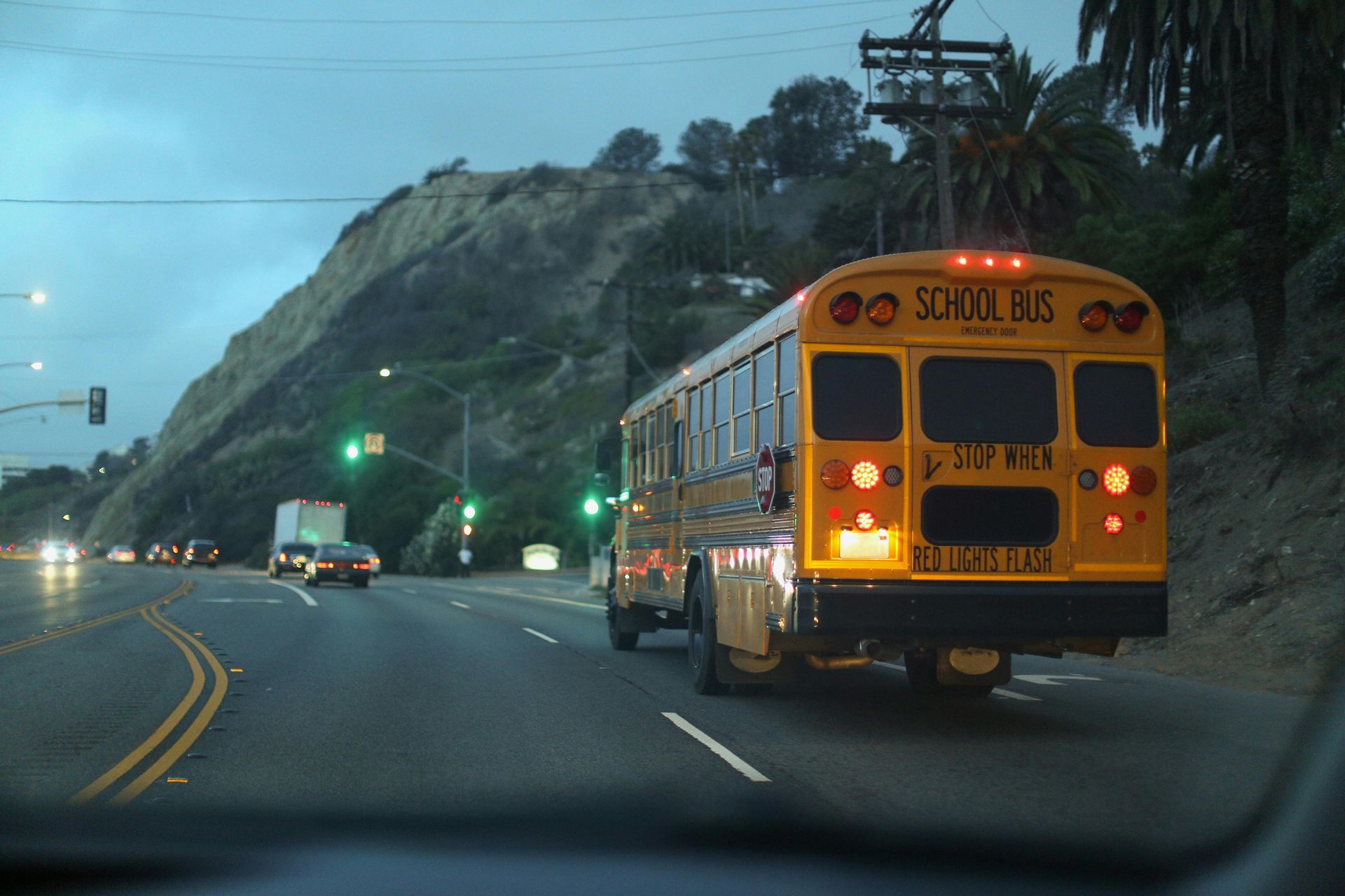 Yellow school bus driving on a road, passing a cliffside, cloudy sky, green traffic lights.