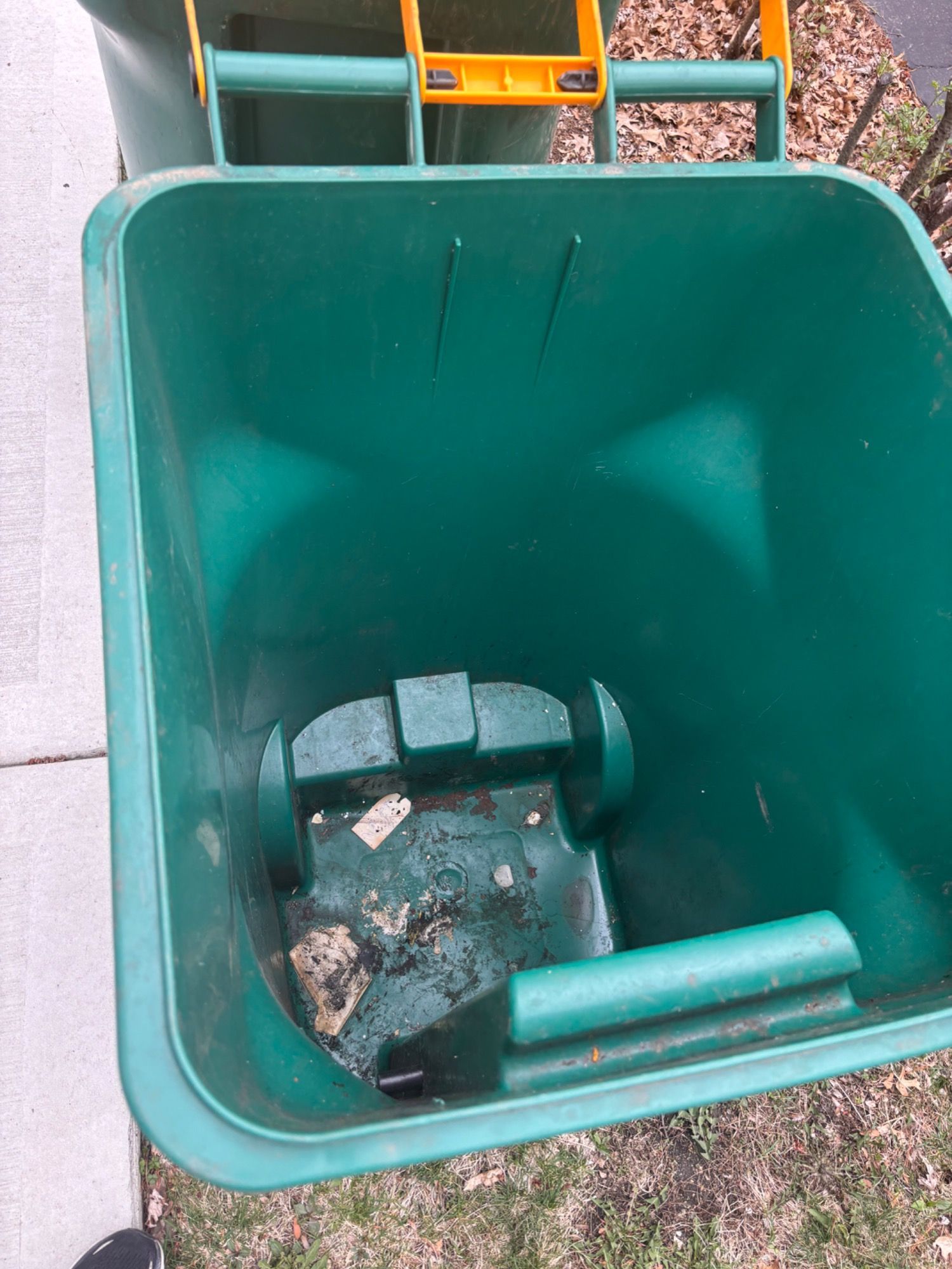 A green trash can with a yellow handle is sitting on the sidewalk.