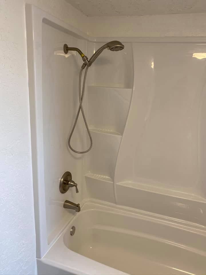 White bathtub and shower with beige walls, built-in shelves, and a handheld showerhead in a bathroom.