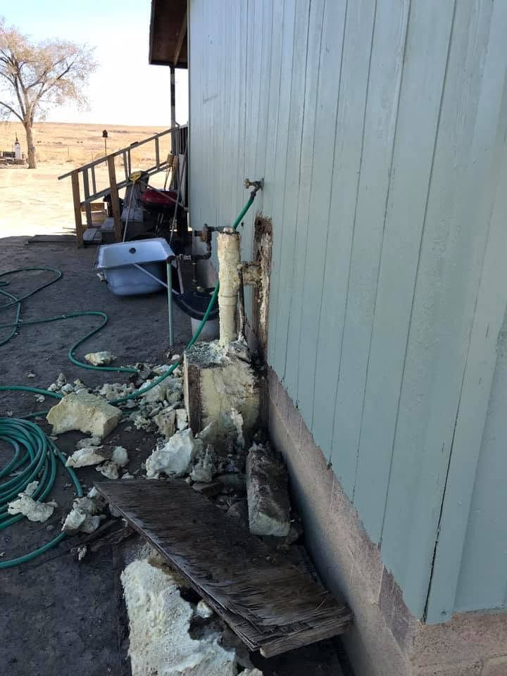 Side of a gray building with a damaged foundation, scattered rocks, hoses, and a small porch area.