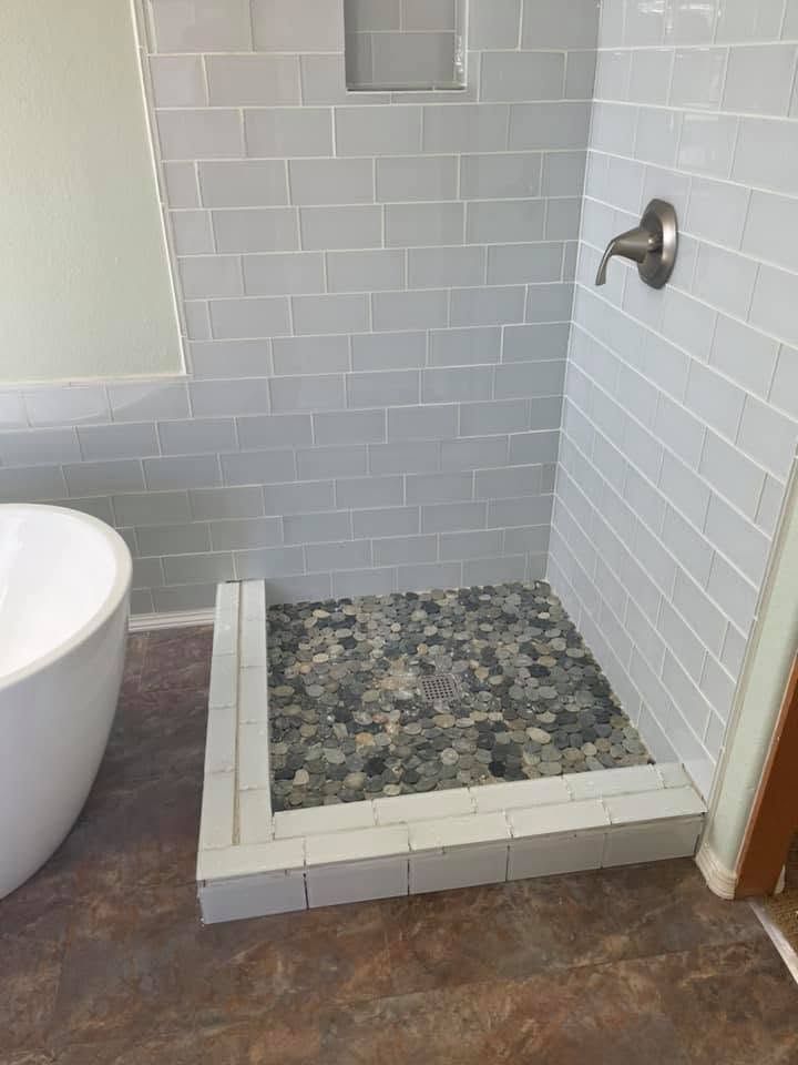 Tiled bathroom shower with pebble floor, white curb, and wall-mounted shower head beside a bathtub