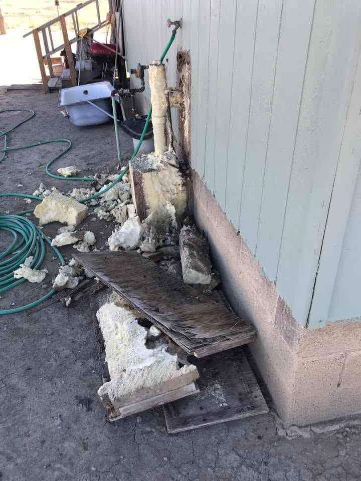 Damaged house exterior with broken boards, debris, and exposed pipes beside a wall