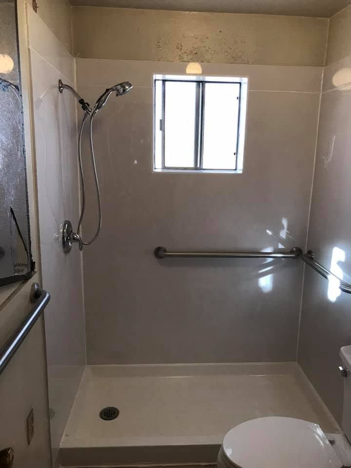 Empty tiled bathroom with a walk-in shower, grab bar, window, and toilet in the foreground