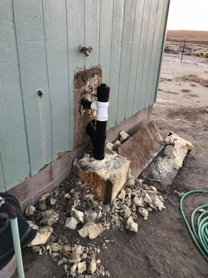 Damaged exterior wall with exposed black pipe and broken concrete near a building foundation
