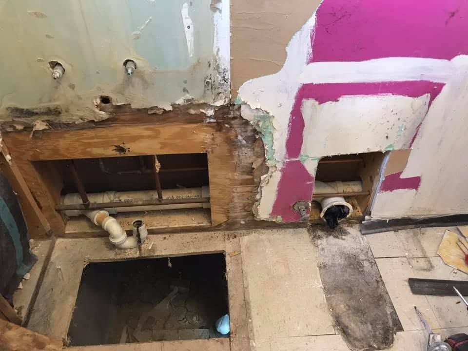 Damaged wall with two open floor holes and exposed pipes in a room under renovation