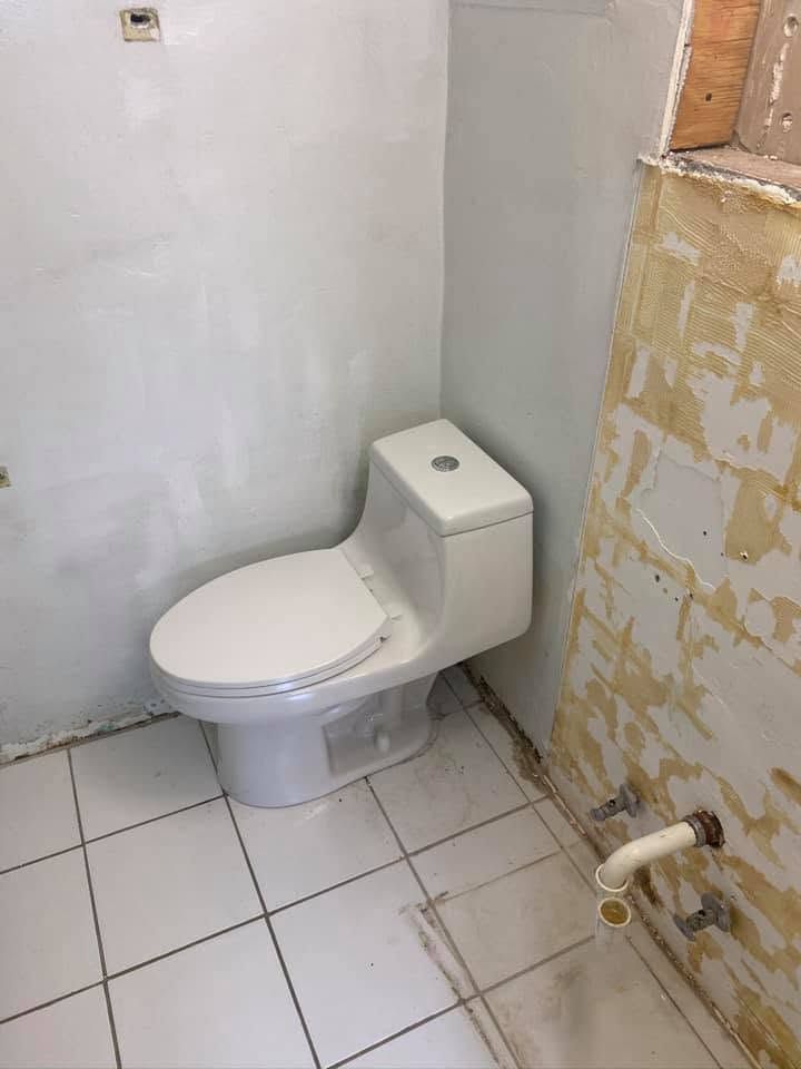 Small white bathroom with a toilet beside a tiled wall and exposed plumbing.