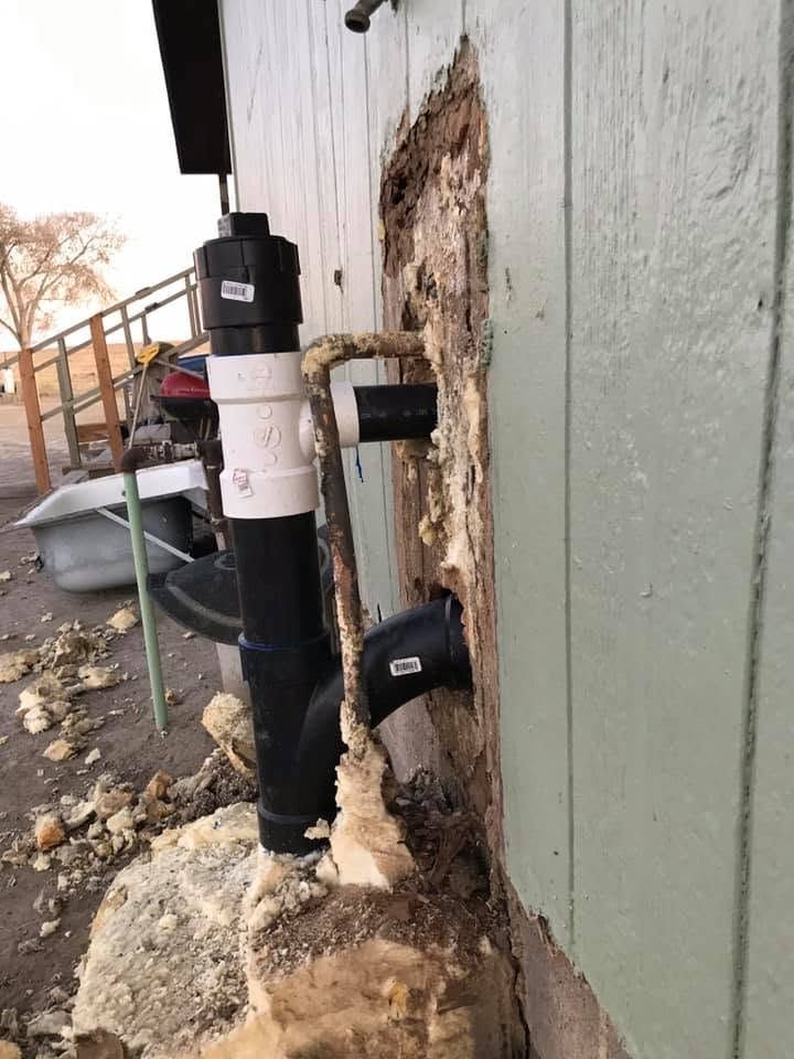 Damaged exterior wall with exposed plumbing pipes and crumbling stucco near a foundation area