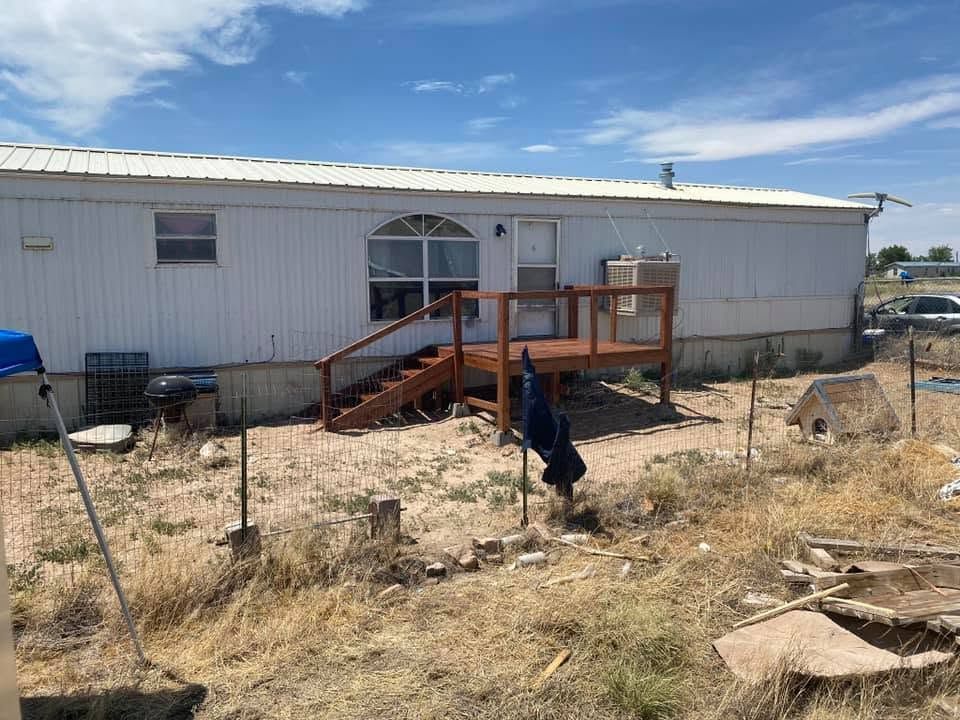 A weathered mobile home with a small wooden porch in a dry, fenced yard.