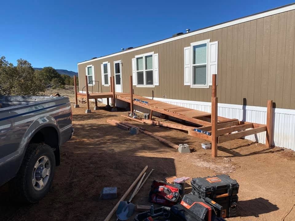 Modular home on a dirt lot with a wooden ramp, partially built deck, and truck parked beside it