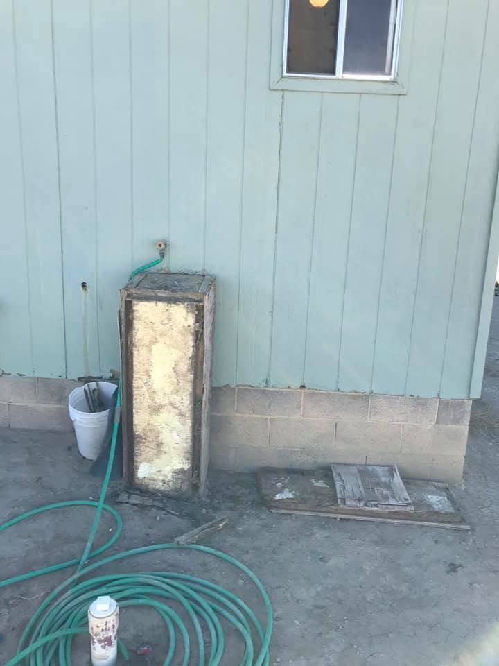 Outdoor utility area with coiled green hose, bucket, and a worn upright unit against a light blue wall.